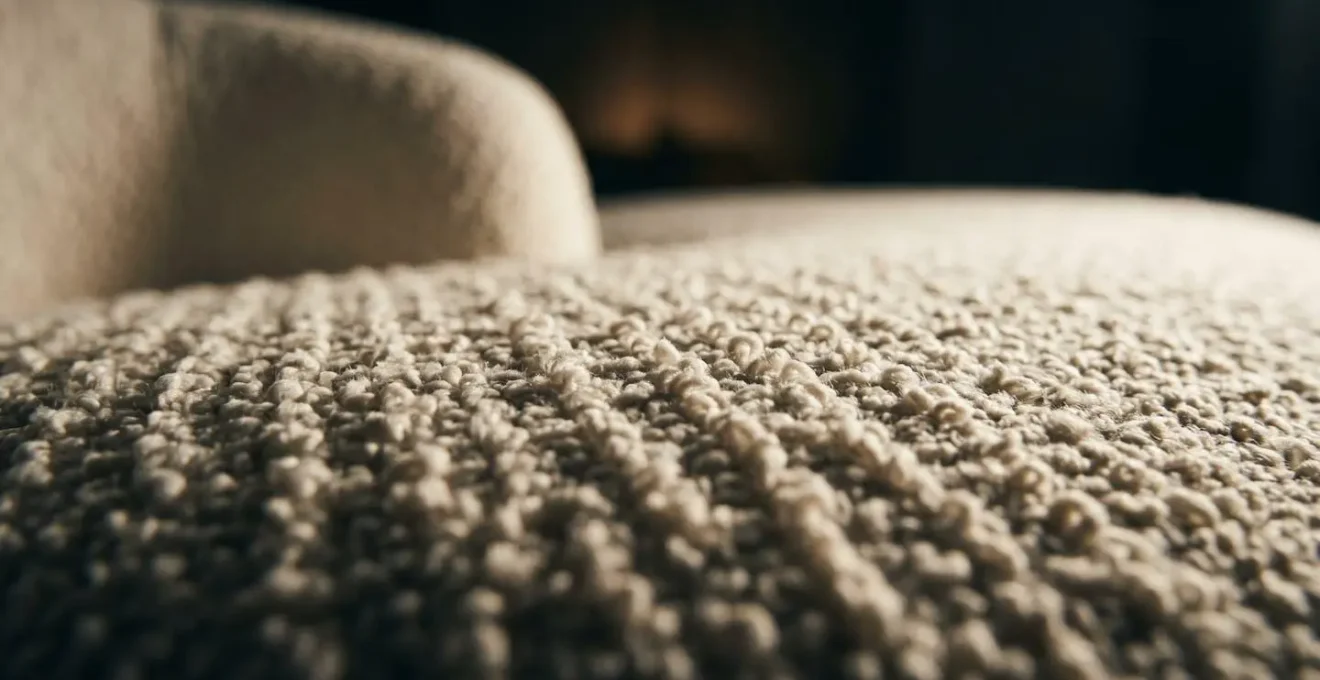 A macro photograph showing the textured surface of a bouclé armchair, with light from a low angle creating deep shadows and highlighting the fabric's tactile quality.
