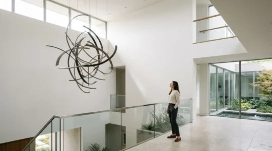 A modern residential atrium with soaring double-height ceiling where a monumental suspended sculpture hangs gracefully, maintaining clear circulation paths below while commanding visual presence